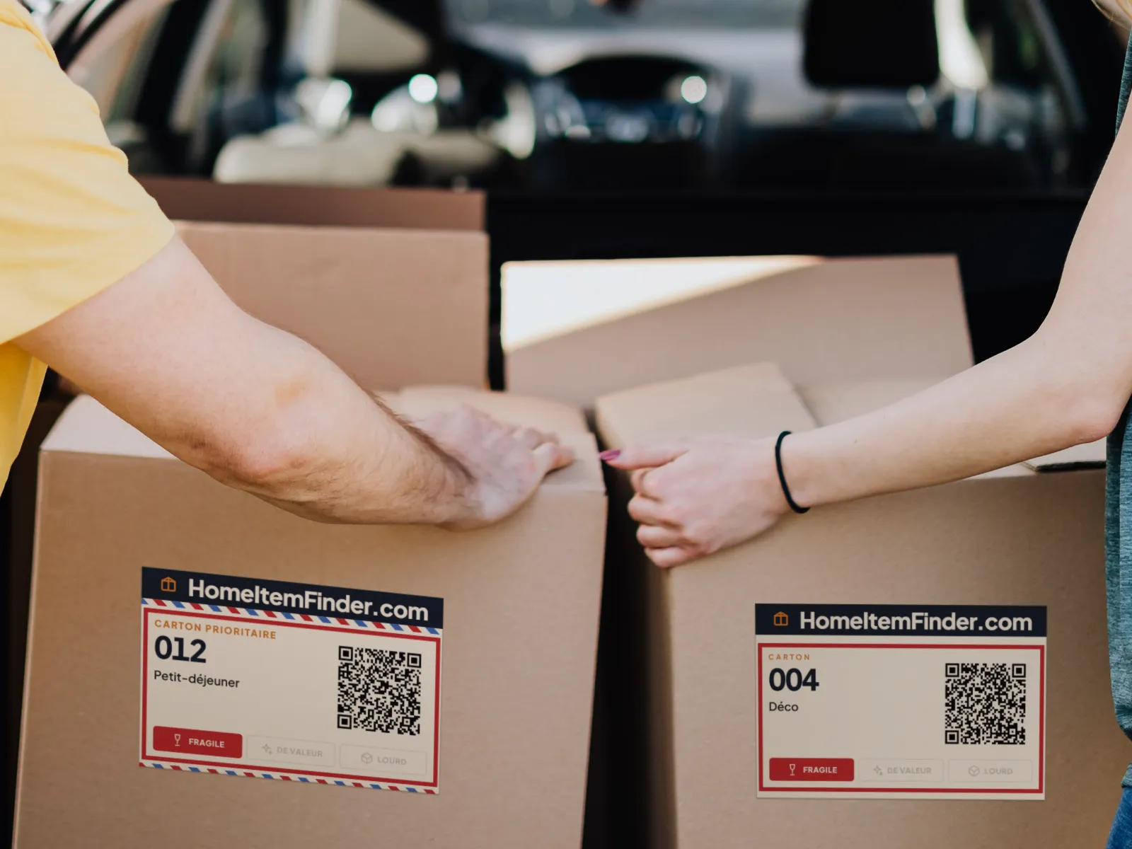 Deux personnes chargent des cartons de déménagement dans une voiture. Les cartons de devant portent les étiquettes de l’app : le 012 et le 004, tous deux marqués fragiles.